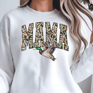 May include: White sweatshirt with the word "MAMA" in camouflage print. A detailed illustration of a flying duck is below the text. The duck has brown, green, and yellow feathers.
