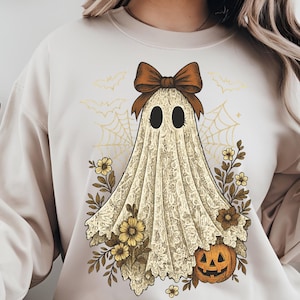 K&ouml;nnte beinhalten: Cremefarbenes Sweatshirt mit einem Geisterdesign im Vintage-Stil. Der Geist tr&auml;gt eine braune Schleife und ist mit einem spitzenartigen Laken drapiert, umgeben von Blumen, einem K&uuml;rbis, Spinnweben und Flederm&auml;usen. Ein festliches Halloween-Themen-Grafik.