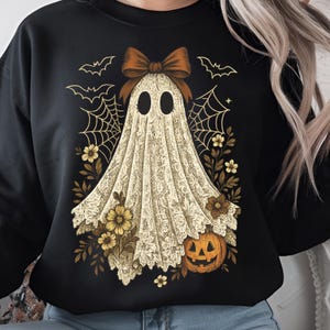 K&ouml;nnte beinhalten: Schwarzes Sweatshirt mit einem Geisterdesign, das eine Schleife, Spinnweben, Blumen, Flederm&auml;use und einen K&uuml;rbis zeigt. Der Geist ist in einem spitzenartigen Stoff drapiert. Ein Halloween-Motiv.