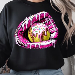 Puede incluir: Una sudadera negra con un gr&aacute;fico rosa y blanco de una pelota de softbol en un guante de receptor con glaseado rosa y chispas.