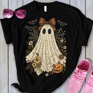 K&ouml;nnte beinhalten: Schwarzes T-Shirt mit einem Halloween-Geisterdesign. Der Geist ist in einem wei&szlig;en, spitzenartigen Stoff drapiert und hat eine braune Schleife. Das Design umfasst Flederm&auml;use, Spinnweben, florale Akzente und einen geschnitzten K&uuml;rbis.