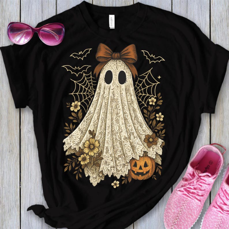 K&ouml;nnte beinhalten: Schwarzes T-Shirt mit einem Halloween-Geisterdesign. Der Geist ist in einem wei&szlig;en, spitzenartigen Stoff drapiert und hat eine braune Schleife. Das Design umfasst Flederm&auml;use, Spinnweben, florale Akzente und einen geschnitzten K&uuml;rbis.