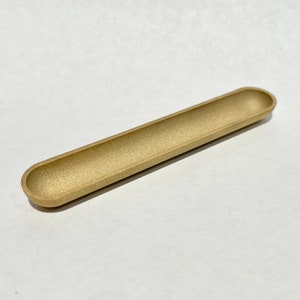 Puede incluir: Un plato de metal largo y estrecho de color dorado con extremos redondeados. El plato está ligeramente curvado y tiene un acabado liso y mate.