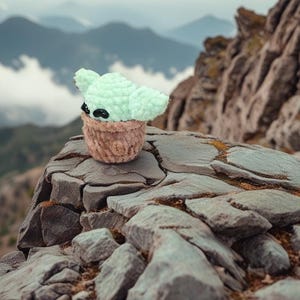 May include: A crocheted Baby Yoda doll with large ears and black eyes, perched on a rocky surface. The doll is light green and brown, resembling the character from the Star Wars series. The background features a mountain range under a cloudy sky.
