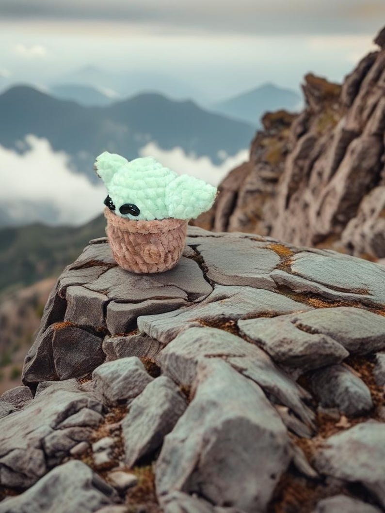 May include: A crocheted Baby Yoda doll with large ears and black eyes, perched on a rocky surface. The doll is light green and brown, resembling the character from the Star Wars series. The background features a mountain range under a cloudy sky.