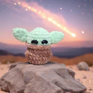 May include: A handmade crochet figure of a character with large ears and black eyes, in a light green color. The figure is perched on a rock in a desert landscape with a starry sky and a shooting star in the background. The figure is a plush toy.