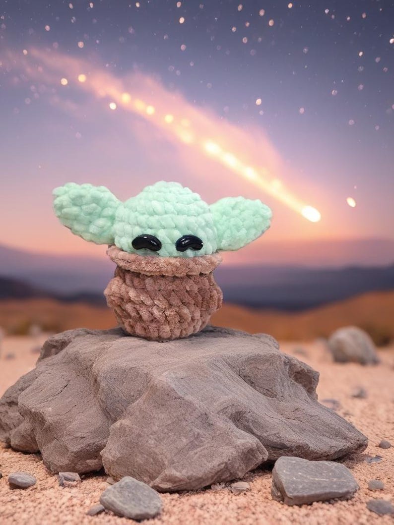 May include: A handmade crochet figure of a character with large ears and black eyes, in a light green color. The figure is perched on a rock in a desert landscape with a starry sky and a shooting star in the background. The figure is a plush toy.