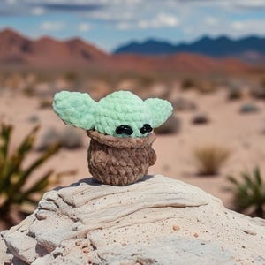 May include: A crocheted figure of a character with large ears and black eyes, in a brown pot. The figure is light green and brown, set against a desert landscape with mountains and a cloudy sky.