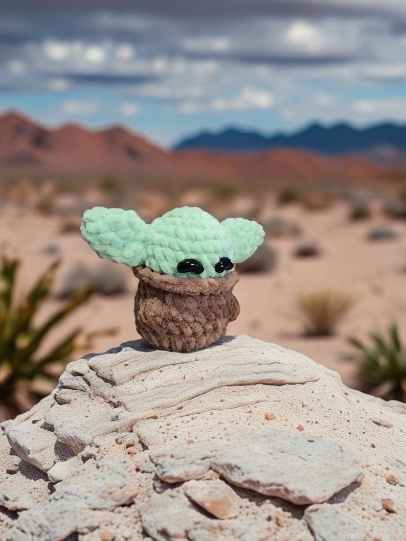 May include: A crocheted figure of a character with large ears and black eyes, in a brown pot. The figure is light green and brown, set against a desert landscape with mountains and a cloudy sky.