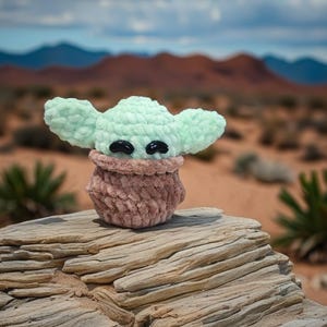 May include: A crocheted doll of a character with large ears and black eyes. The doll has a light green head and ears, a brown body, and is sitting on a weathered rock. The background features a desert landscape with mountains and a cloudy sky.