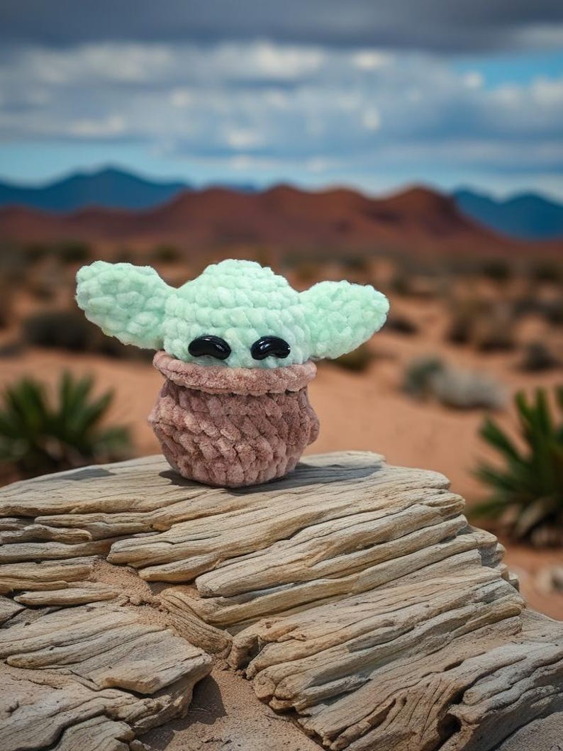 May include: A crocheted doll of a character with large ears and black eyes. The doll has a light green head and ears, a brown body, and is sitting on a weathered rock. The background features a desert landscape with mountains and a cloudy sky.