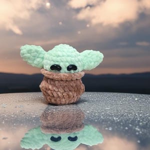 May include: A handmade crochet doll in the likeness of a popular character. The doll features large, mint green ears and a brown body. Black, oval-shaped eyes are visible. The doll is reflected in the shiny surface below, with a cloudy sky in the background.