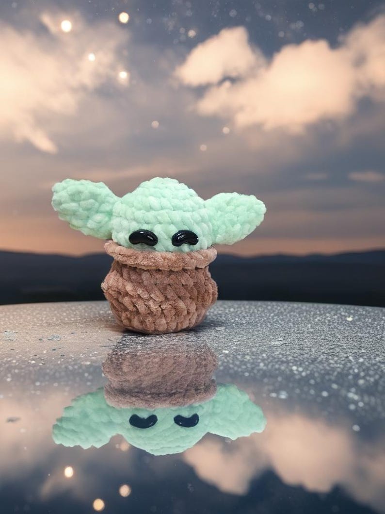 May include: A handmade crochet doll in the likeness of a popular character. The doll features large, mint green ears and a brown body. Black, oval-shaped eyes are visible. The doll is reflected in the shiny surface below, with a cloudy sky in the background.