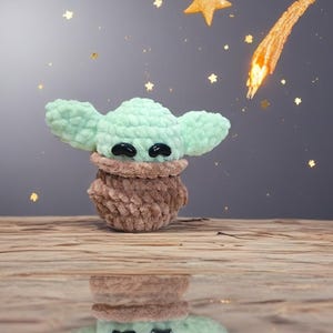 May include: A handmade crochet figure of a character with large ears, in mint green and brown. The figure has black eyes and is set against a starry background with a shooting star. The figure is reflected in a glossy surface.