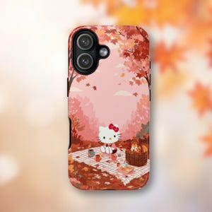 May include: A phone case featuring a cartoon picnic scene with Hello Kitty. The case has a pink and red autumn-themed background with falling leaves and a picnic basket. The case is designed to fit a smartphone.