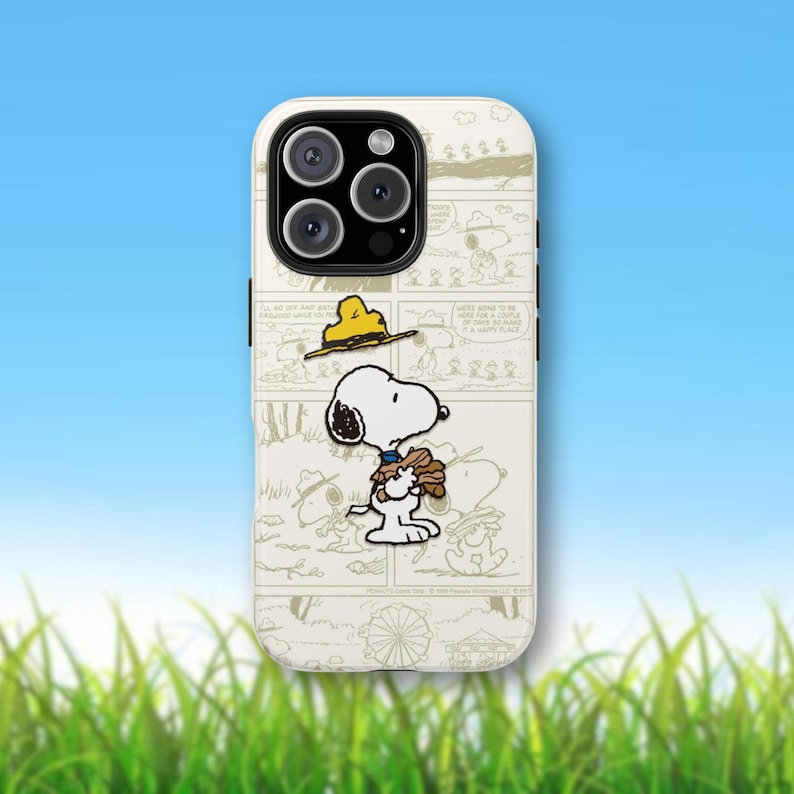 May include: A white phone case featuring a comic strip design with Snoopy and Woodstock characters. The case has a cream-colored background with a blue sky and green grass in the background.
