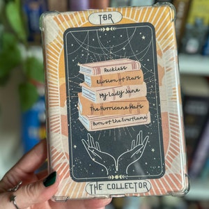 Puede incluir: Una tarjeta de plástico transparente con fondo negro y una pila de libros con los títulos "Reckless", "Illusion of Stars", "My Lady Jane", "The Hurricane Wars" y "Burn of the Everflame". La tarjeta tiene un borde dorado y el texto "TBR" y "THE COLLECTOR" en dorado.