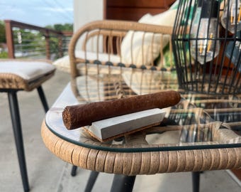 May include: A brown cigar resting on a white rectangular ashtray on a glass-top table with a wicker base. The table is in front of a wicker chair with a white cushion.
