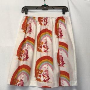 May include: A white skirt with a rainbow and Care Bear print. The Care Bears are holding hearts and are kissing. The skirt has a gathered waist.