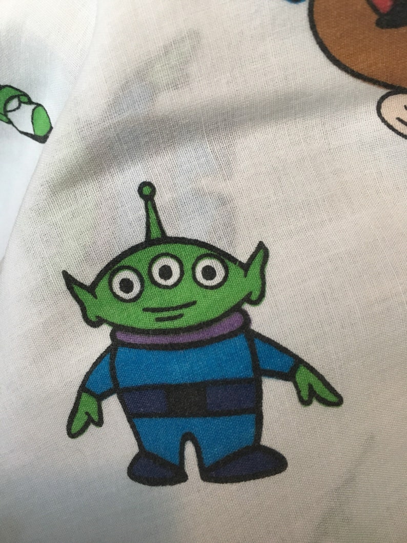 Op de afbeelding: Een groene alien met drie ogen en een paarse kraag draagt een blauw pak. De alien staat op een witte achtergrond.