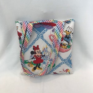May include: A white tote bag with a rainbow striped handle. The bag features a colorful print of Mickey Mouse, Minnie Mouse, Donald Duck, and the Cinderella Castle.