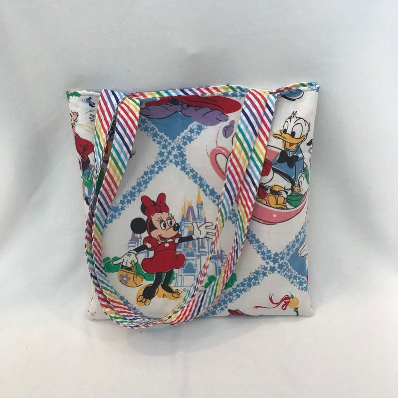 May include: A white tote bag with a rainbow striped handle. The bag features a colorful print of Mickey Mouse, Minnie Mouse, Donald Duck, and the Cinderella Castle.
