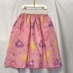May include: A pink skirt with a Care Bears print. The skirt has a white elastic waistband and features yellow, purple, and pink Care Bears with butterflies and flowers.