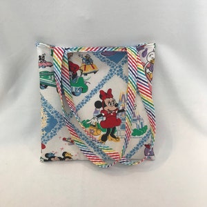 May include: A white canvas tote bag with rainbow striped straps. The bag features a colorful print of Mickey Mouse and Minnie Mouse characters in a whimsical setting. The print includes a castle, cars, and stars.
