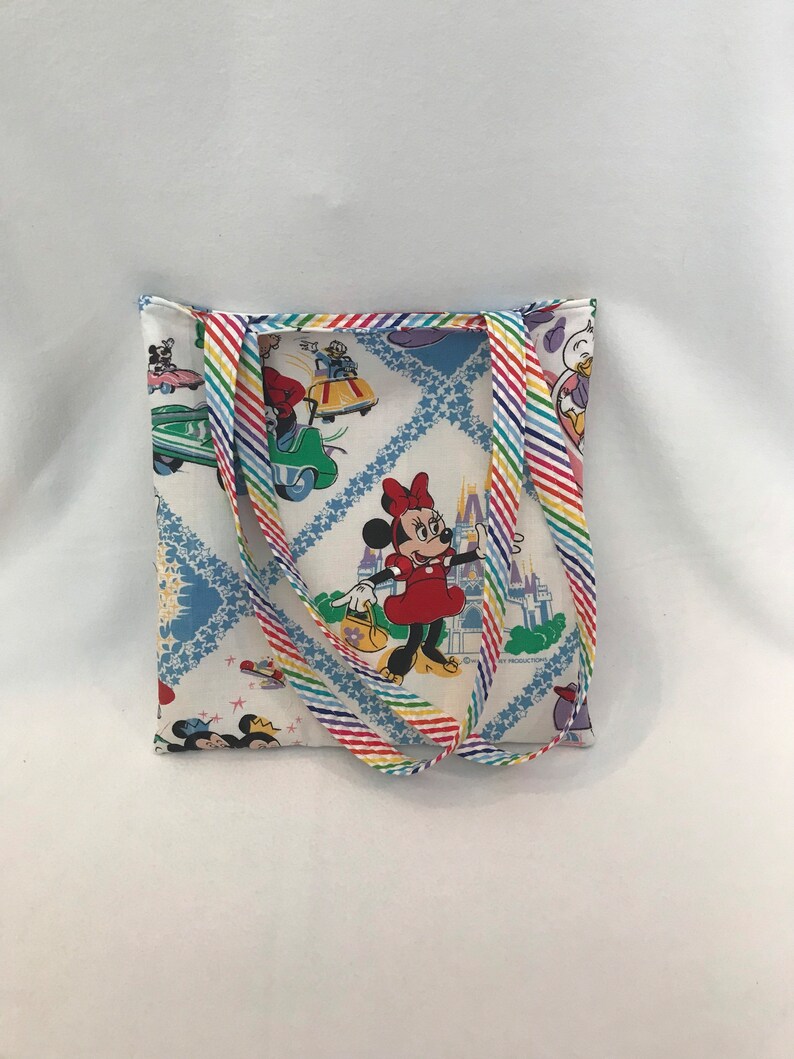 May include: A white canvas tote bag with rainbow striped straps. The bag features a colorful print of Mickey Mouse and Minnie Mouse characters in a whimsical setting. The print includes a castle, cars, and stars.