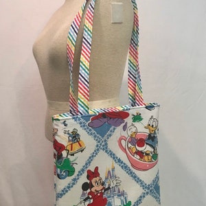 May include: A white canvas tote bag with a colorful rainbow striped strap. The bag features a vintage Disney print with Mickey Mouse, Minnie Mouse, Donald Duck, and the Cinderella Castle. The print is on a white background with blue and white diamond shapes.