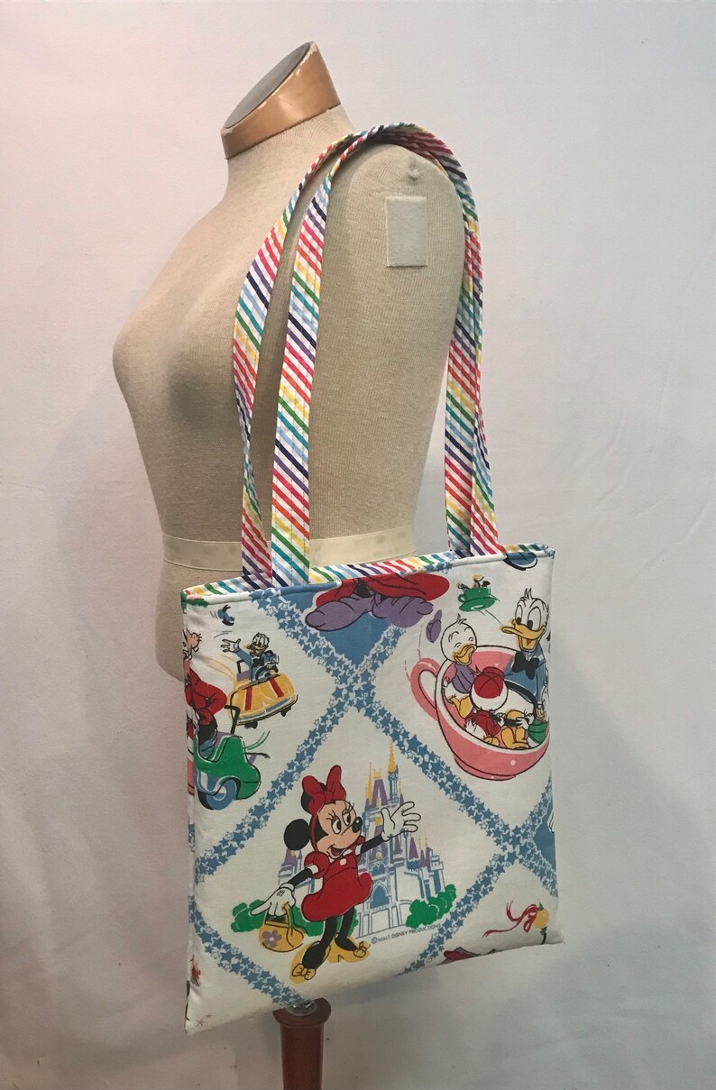 May include: A white canvas tote bag with a colorful rainbow striped strap. The bag features a vintage Disney print with Mickey Mouse, Minnie Mouse, Donald Duck, and the Cinderella Castle. The print is on a white background with blue and white diamond shapes.