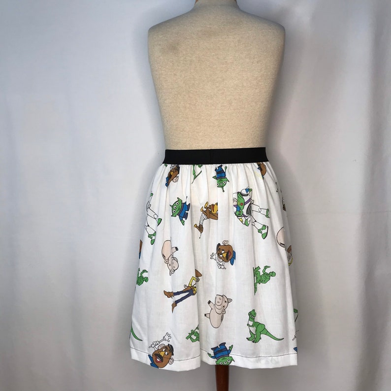 Op de afbeelding: Een witte rok met een zwarte tailleband en een patroon van Toy Story-personages. De personages zijn Woody, Buzz Lightyear, Rex en Mr. Potato Head.