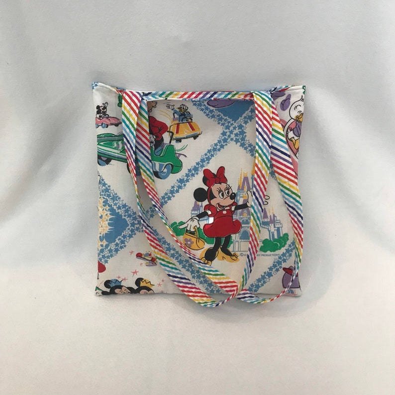 May include: A white canvas tote bag with rainbow striped straps. The bag features a colorful print of Mickey Mouse and Minnie Mouse characters, including the Disney castle.