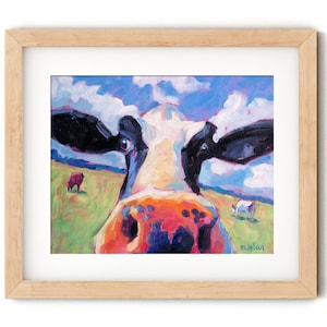 May include: Framed art print of a cow's face, painted in vibrant colors. The cow's nose is orange and pink, with black and white markings. The background shows a green field, blue sky, and other cows. The artwork is in a light wood frame.