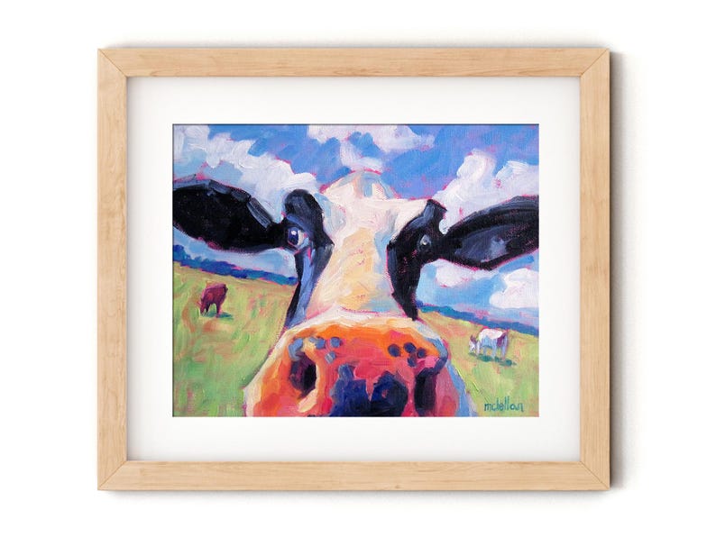 May include: Framed art print of a cow's face, painted in vibrant colors. The cow's nose is orange and pink, with black and white markings. The background shows a green field, blue sky, and other cows. The artwork is in a light wood frame.