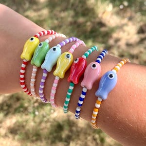 May include: A collection of colorful beaded bracelets, each featuring a unique fish-shaped charm. The bracelets showcase a variety of colors, including yellow, green, blue, red, and pink, with coordinating bead colors. The bracelets are worn on a wrist.
