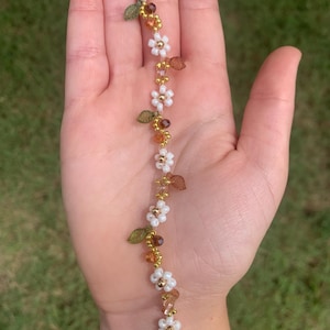 May include: A delicate floral bracelet featuring white daisy-like beads, gold accents, and small leaf details. The bracelet is strung on a gold chain and has a gold clasp.
