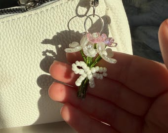 May include: A beaded flower bouquet keychain with white and pink petals, green stems, and a silver clasp. The keychain is attached to a white leather bag with a zipper.