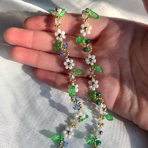 May include: Two beaded floral chain accessories. Each chain features white flower beads, green leaf beads, and blue and green crystal beads. The chains are connected by a gold-colored chain.
