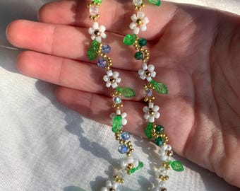 May include: Two beaded floral chain accessories. Each chain features white flower beads, green leaf beads, and blue and green crystal beads. The chains are connected by a gold-colored chain.