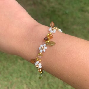 May include: A beaded bracelet featuring white flower accents, gold beads, and brown leaf details. The bracelet is adorned with small, clear beads and is designed to be worn around the wrist.