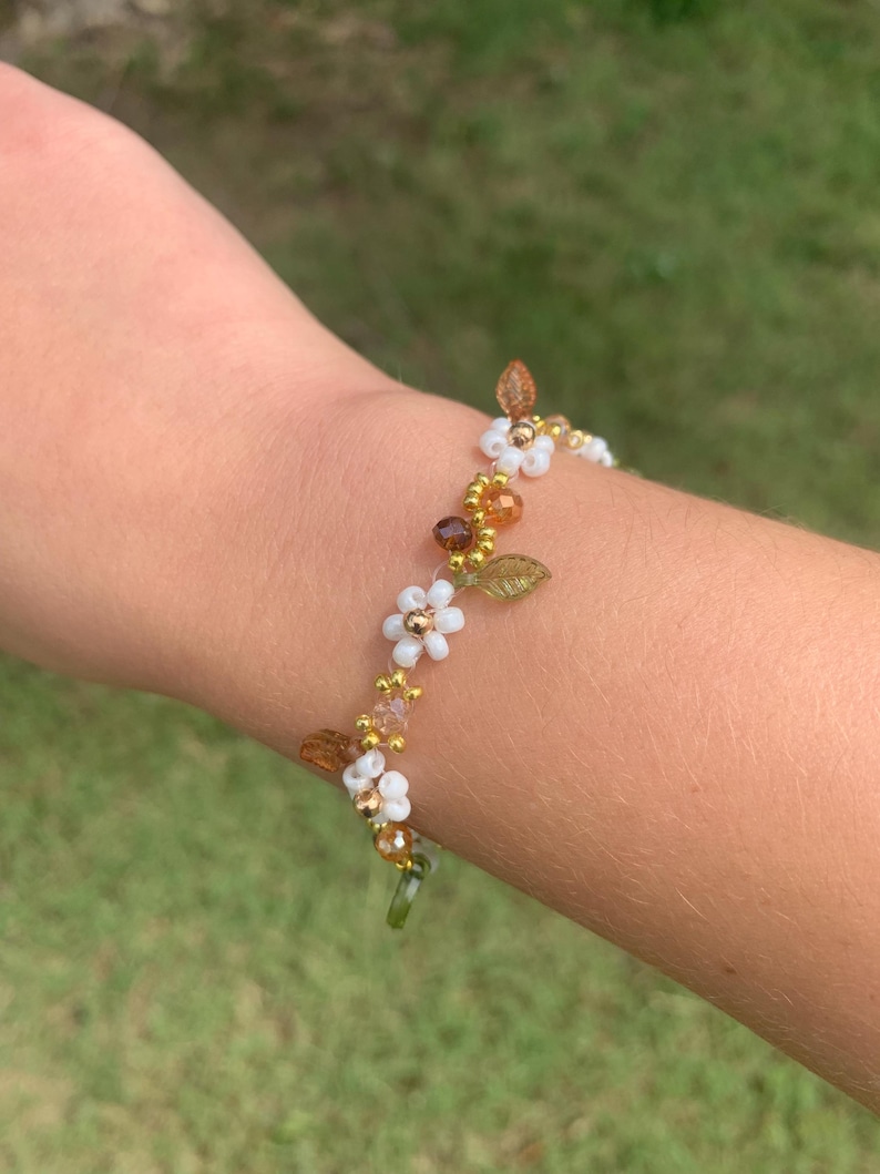 May include: A beaded bracelet featuring white flower accents, gold beads, and brown leaf details. The bracelet is adorned with small, clear beads and is designed to be worn around the wrist.