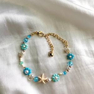 May include: A beaded bracelet featuring a gold chain and clasp. The bracelet is adorned with light blue flower beads, white star beads, and a beige starfish bead. Additional beads are white, gold, and clear.