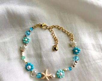 Può includere: Un braccialetto di perline con catena e chiusura dorate. È adornato con perline a forma di fiore azzurro, perline a stella bianche e una perlina a forma di stella marina beige. Il braccialetto include anche perline bianche, dorate e trasparenti.
