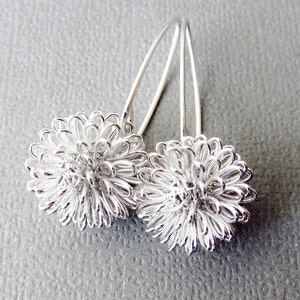 May include: A pair of silver-colored earrings. Each earring features a spherical, wire-like design resembling a flower. The earrings hang from curved, thin silver hooks. The background is a solid, light gray color.
