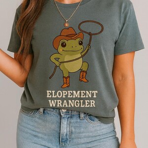Könnte beinhalten: Grünes T-Shirt mit dem Text "ELOPEMENT WRANGLER". Das Shirt zeigt einen Cartoon-Frosch mit Cowboyhut und Stiefeln, der ein Lasso hält. Der Frosch ist grün, der Hut und die Stiefel sind braun.