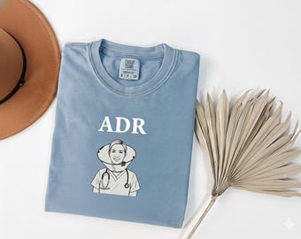 May include: A light blue t-shirt with the letters "ADR" above a black and white illustration of a person wearing a medical collar and stethoscope. The shirt is made of a soft material.