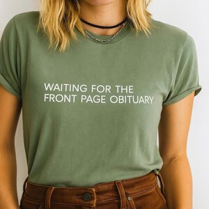 Puede incluir: Camiseta verde oliva con el texto blanco "WAITING FOR THE FRONT PAGE OBITUARY". La camiseta se combina con pantalones marrones. La modelo lleva un choker negro y un collar de cadena plateada.