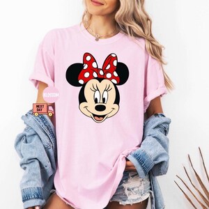 May include: A light pink t-shirt featuring a large graphic of Minnie Mouse's face. Minnie Mouse has black ears, a red bow with white polka dots, and a smiling expression. The shirt is part of a casual wear collection.