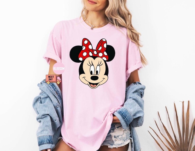 May include: A light pink t-shirt featuring a large graphic of Minnie Mouse's face. Minnie Mouse has black ears, a red bow with white polka dots, and a smiling expression. The shirt is part of a casual wear collection.
